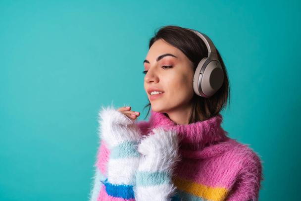 Fiatal nő fényes sokszínű pulóverben kék háttérrel nagy zajszűrő fejhallgatóval, élvezi kedvenc zenéjét, zeneszerető - Fotó, kép