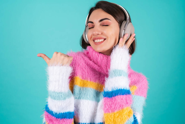 Junge Frau in hellem, buntem Pullover vor blauem Hintergrund mit großen, geräuschunterdrückenden Kopfhörern, genießt ihre Lieblingsmusik, zeigt mit dem Finger nach links - Foto, Bild