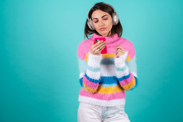 Young woman in a bright multicolored sweater on a blue background in large noise canceling headphones holds a phone with a bored displeased look - Photo, Image