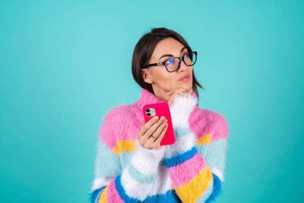 Young woman in a bright multicolored sweater on a blue background with glasses, holding a phone, pensive, dreamy looking to the side - Foto, Imagem