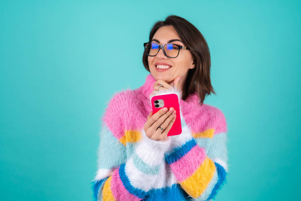 眼鏡をかけた青い背景に明るいマルチカラーのセーターを着た若い女性が電話を持って、熱心に笑う - 写真・画像