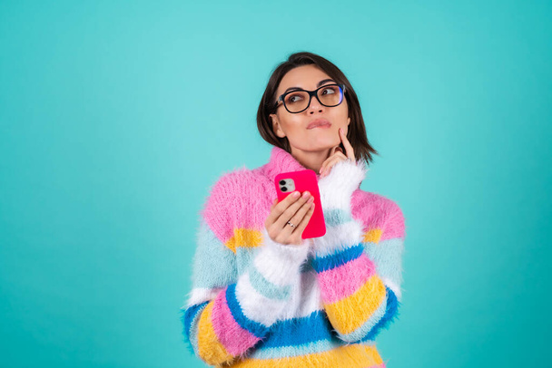 Jeune femme dans un pull multicolore lumineux sur un fond bleu avec des lunettes, tenant un téléphone, cher, rêveur regardant vers le côté - Photo, image