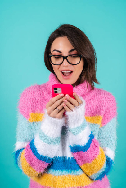 A young woman in a bright multi-colored sweater on a blue background with glasses, holds a phone, looks at the screen in surprise with an open mouth - Fotografie, Obrázek