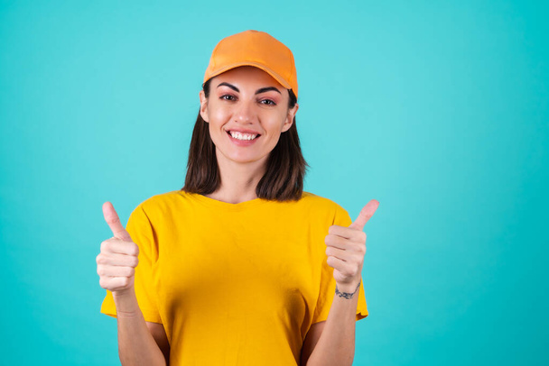 Young woman delivery service worker, courier, in a cap on a blue background smiles broadly and shows thumbs up - Photo, Image