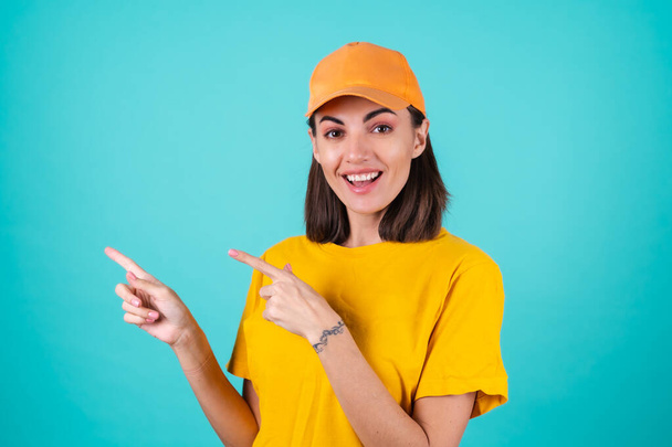 Trabajadora de servicio de entrega joven, mensajera, con una gorra sobre un fondo azul sonríe ampliamente y señala los dedos a la izquierda - Foto, imagen