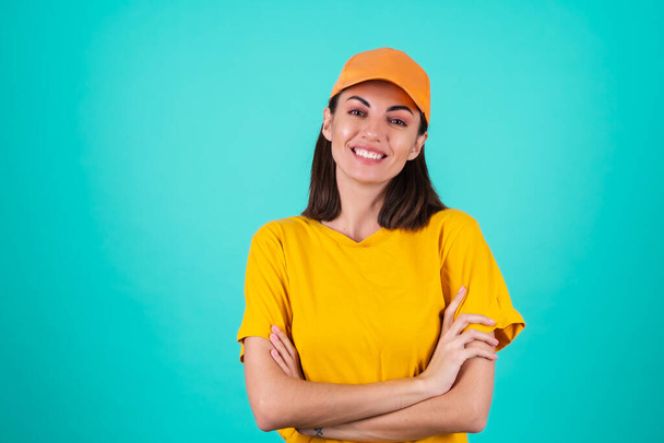 Genç kadın teslimatçı, kurye, mavi arka planda turuncu sarı şapkalı, geniş bir gülümseme ve çapraz kollarla duruyor. - Fotoğraf, Görsel