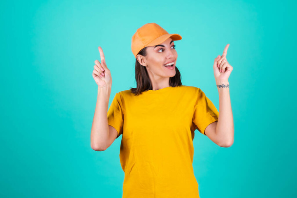 Trabajadora de servicio de entrega joven, mensajera, en una gorra sobre un fondo azul sonríe ampliamente y señala con los dedos hacia arriba - Foto, Imagen