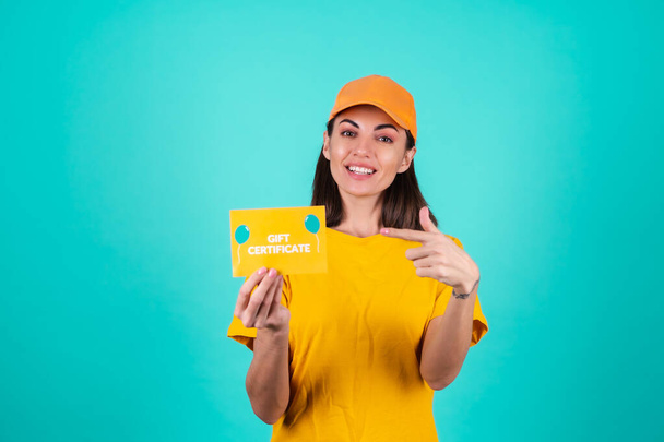 Młoda kobieta doręczyciel, kurier, w pomarańczowej żółtej czapce na niebieskim tle posiada bon podarunkowy i uśmiecha się szeroko - Zdjęcie, obraz