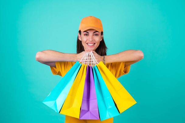 Young woman delivery service worker, courier, in an orange yellow cap on a blue background, holds multicolored shopping paper bags, delivers an order - Fotoğraf, Görsel