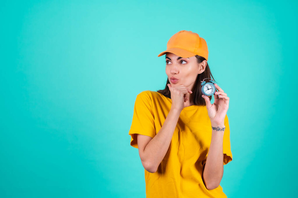 Jeune femme livreuse de services, courrier, dans une casquette jaune orange sur un fond bleu tient une horloge et regarde pensivement sur le côté - Photo, image