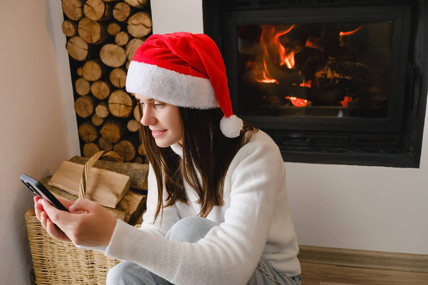 Close up of smiling young woman celebrate Christmas at home text message greeting on cellphone. Happy millennial female have smartphone call near fireplace on winter holidays. Communication concept - Foto, imagen