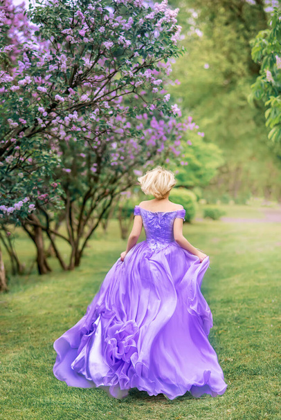 girl in a long lilac, blue dress runs in the park. Photo from the back. Lilac flowers grow in the park - Photo, Image