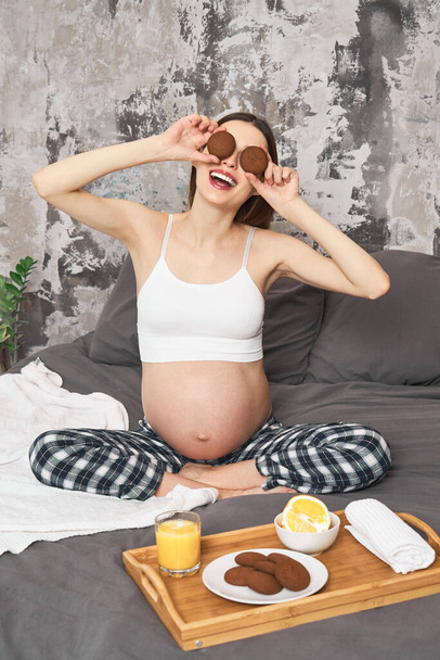 Brunch time, young pregnant woman in pajamas, drinking orange juice, eating cookies, sitting on bed, having good time. The concept of motherhood, parenting, childbirth, nutrition of a pregnant woman. - Photo, image