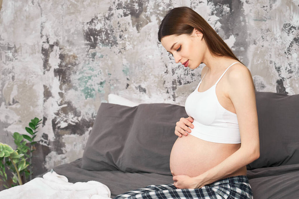 Pregnant woman feeling happy at home while taking care of her child. The young expecting mother holding baby in pregnant belly. Maternity prenatal care and woman pregnancy concept. - 写真・画像