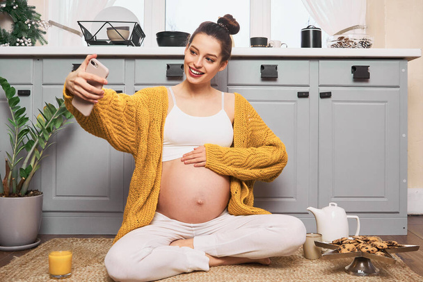 Young Caucasian pregnant girl in a yellow cardigan with a naked belly, sitting on the kitchen floor and taking a selfie, taking pictures of her belly, video calling. Pregnancy and motherhood concept. - Foto, imagen