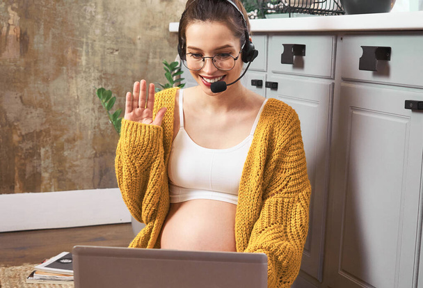 Close-up, a cute attractive young pregnant woman in a yellow cardigan with a naked belly, glasses and a headset, sits on the kitchen floor and works remotely, conducts a video conference. - Foto, Imagen