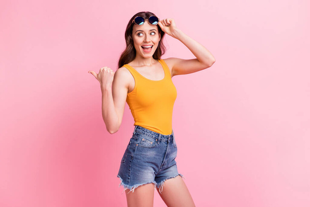 Photo of excited pretty young woman wear yellow singlet dark eyewear pointing thumb up empty space isolated pink color background - Foto, immagini