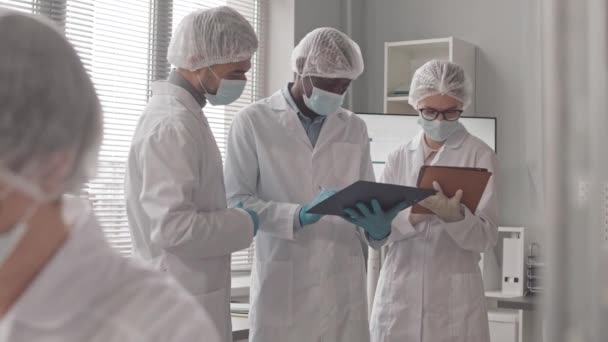 Médium de cientistas caucasianos biraciais e afro-americanos e femininos masculinos vestindo uniforme de trabalho em pé à janela em laboratório, conversando - Filmagem, Vídeo