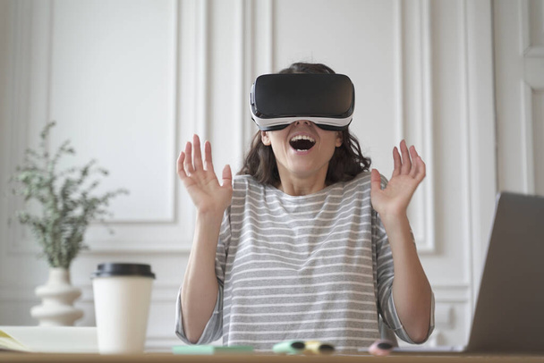 Ενθουσιασμένη επιχειρηματίας κυρία δοκιμές τεχνολογία εικονικής πραγματικότητας φορώντας γυαλιά VR κάθεται στο γραφείο στο σπίτι - Φωτογραφία, εικόνα