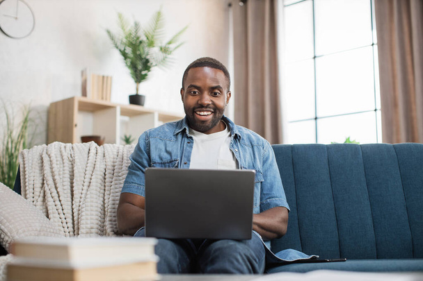 Happy african man sitting on couch and using laptop - Fotó, kép