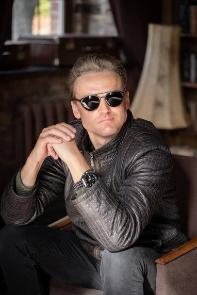 Sexy Blond Man in Stylish Sunglasses and a Leather Jacket Sits in an Armchair with Folded Hands. Close-up Portrait. Vintage Interior Background - Фото, изображение