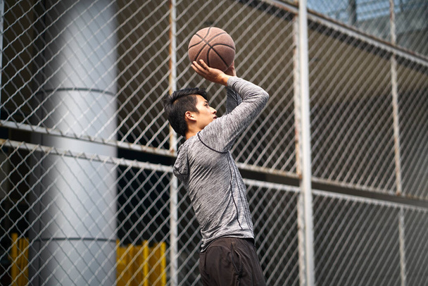 mladý asijský mužský basketbalista dělat skok výstřel na oplocený venkovní soud - Fotografie, Obrázek