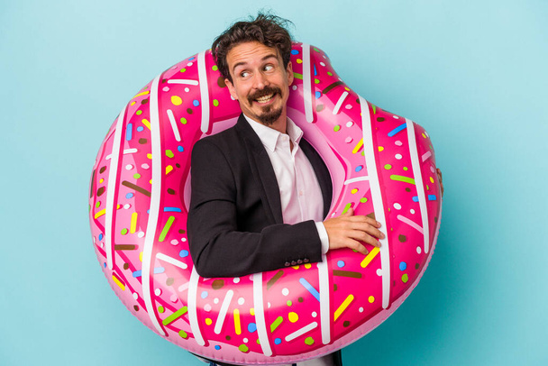 Young business man with inflatable donut isolated on blue background looks aside smiling, cheerful and pleasant. - Photo, Image