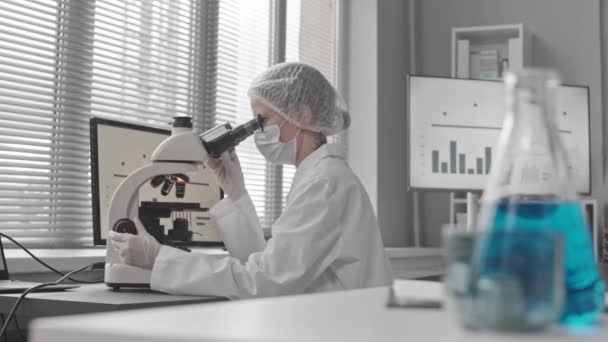 Rastreamento foco de copos com líquidos para jovem cientista caucasiano vestindo uniforme médico e óculos, sentado na mesa em laboratório, usando microscópio, digitando no computador - Filmagem, Vídeo