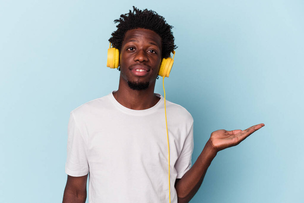 Joven afroamericano escuchando música aislada sobre fondo azul mostrando un espacio de copia en una palma y sosteniendo otra mano en la cintura. - Foto, imagen