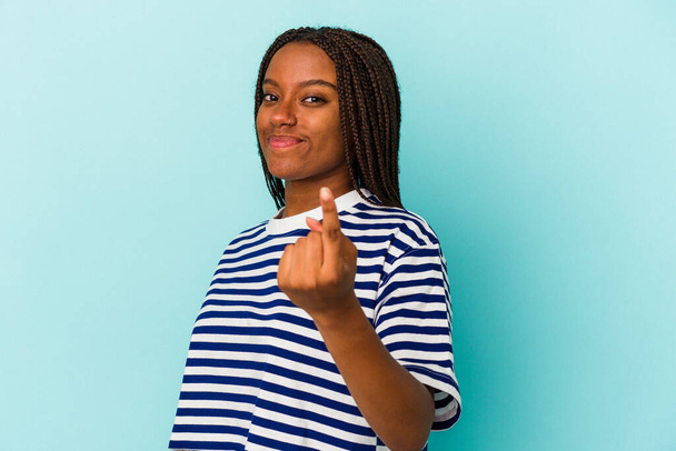 Молода афроамериканська жінка, ізольована на синьому фоні, вказує пальцем на вас, ніби запрошуючи підійти ближче.. - Фото, зображення