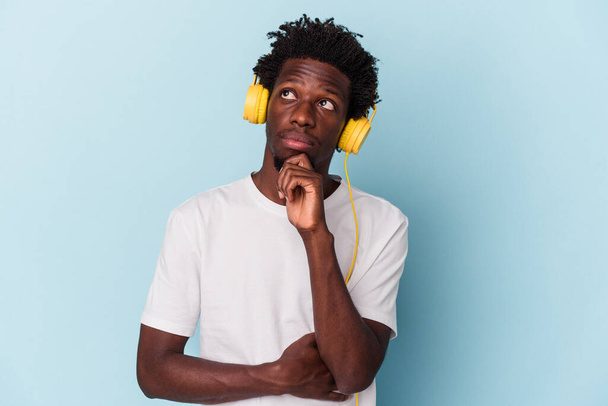 Junger afrikanisch-amerikanischer Mann hört isoliert Musik auf blauem Hintergrund und blickt mit zweifelndem und skeptischem Gesichtsausdruck zur Seite. - Foto, Bild