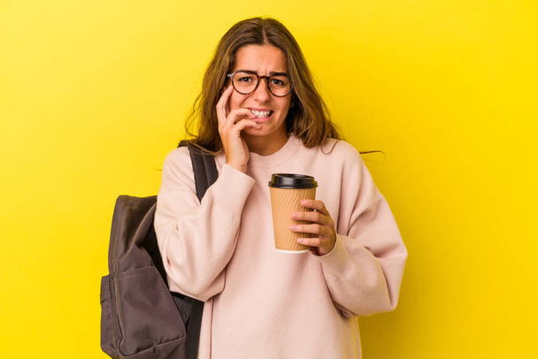 Fiatal kaukázusi diák nő kezében kávé elszigetelt sárga háttér harapás körmök, ideges és nagyon ideges. - Fotó, kép