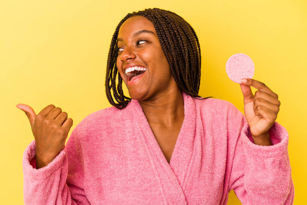 Fiatal afro-amerikai nő fürdőköpenyt visel, kezében egy smink lemez elszigetelt sárga háttér pontokon hüvelykujjával el, nevetve és gondtalanul. - Fotó, kép
