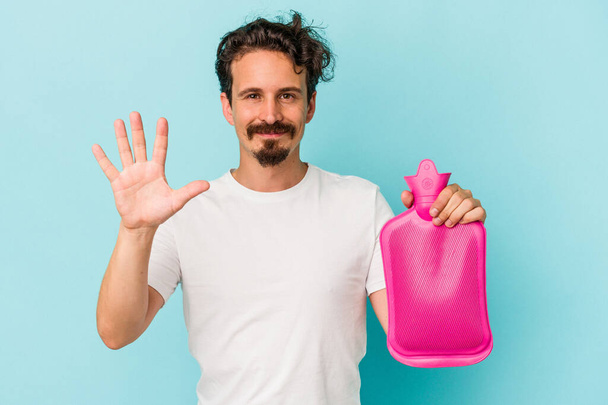 Young caucasian man holding a water bag isolated on blue background smiling cheerful showing number five with fingers. - Photo, Image