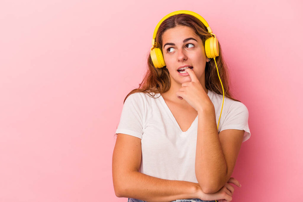 Mladá běloška, která poslouchá hudbu izolovanou na růžovém pozadí, uvolněně přemýšlí o něčem, co se dívá na kopírovací prostor. - Fotografie, Obrázek