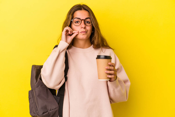 Young caucasian student woman holding coffee isolated on yellow background  with fingers on lips keeping a secret. - Fotografie, Obrázek