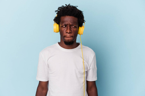 Jeune homme afro-américain à l'écoute de musique isolée sur fond bleu confus, se sent douteux et incertain. - Photo, image