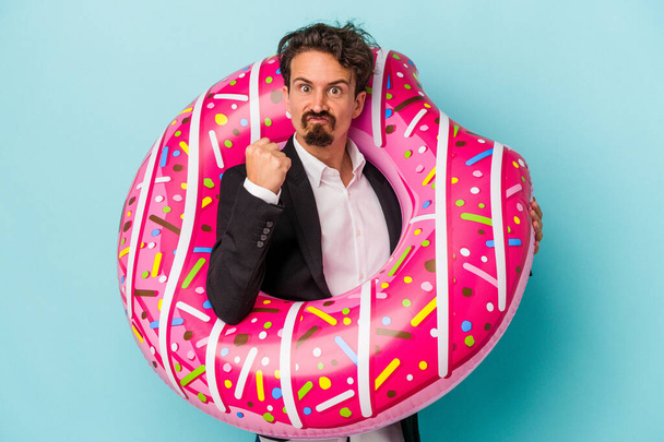 Joven hombre de negocios con donut inflable aislado sobre fondo azul mostrando puño a cámara, expresión facial agresiva. - Foto, imagen