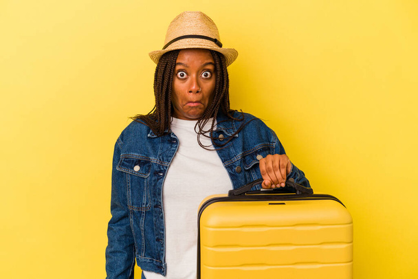 Joven mujer viajera afroamericana sosteniendo maleta aislada sobre fondo amarillo encoge hombros y ojos abiertos confundidos. - Foto, Imagen