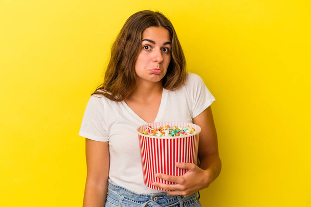 Jonge blanke vrouw met een popcorn geïsoleerd op gele achtergrond haalt schouders en open ogen verward. - Foto, afbeelding