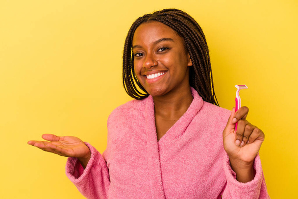 Fiatal afro-amerikai nő fürdőköpenyt visel, kezében egy borotvapengével, sárga alapon elszigetelve, egy tenyéren lévő fénymásoló helyet mutat, és egy másik kezet tart a derekán.. - Fotó, kép