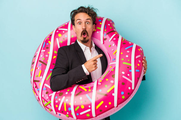 Jovem homem de negócios com donut inflável isolado no fundo azul apontando para o lado - Foto, Imagem