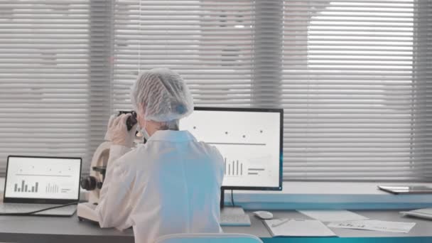 Οπίσθια όψη γυναίκας καυκάσιας ιατρικής ερευνήτριας που φοράει ιατρική ρόμπα και καπέλο, κάθεται στο γραφείο από το παράθυρο στο εργαστήριο, χρησιμοποιώντας μικροσκόπιο υπολογιστή - Πλάνα, βίντεο