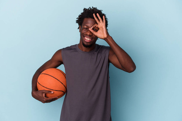 Giovane uomo afro-americano che gioca a basket isolato su sfondo blu eccitato mantenendo ok gesto sugli occhi. - Foto, immagini