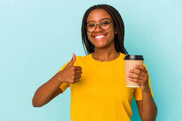 Mladá Afroameričanka drží odnést kávu izolované na modrém pozadí s úsměvem a zvednutím palce nahoru - Fotografie, Obrázek