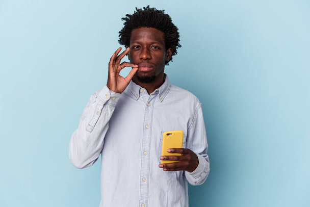 Giovane uomo afroamericano in possesso di cellulare isolato su sfondo blu con le dita sulle labbra mantenendo un segreto. - Foto, immagini