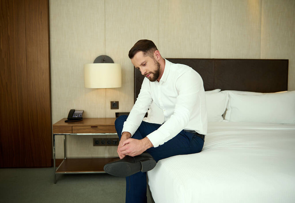 Attractive European middle aged man, successful businessman on a business trip, sits on the edge of the bed in a hotel bedroom and ties the laces on his shoes - Фото, изображение