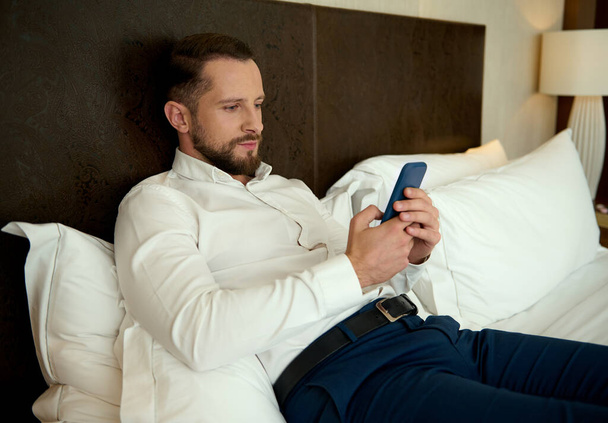 Handsome young European bearded man, successful businessman browsing and chatting on mobile phone, lying on a bed in the bedchamber of a hotel, relaxing after hard day at work and business meetings - Fotografie, Obrázek
