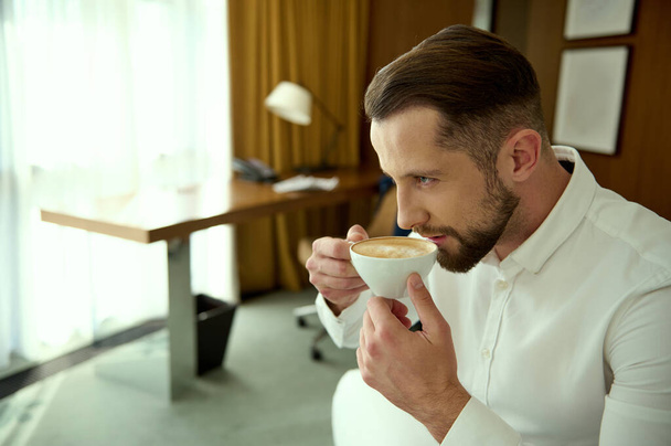 Zelfverzekerde aantrekkelijke knappe jonge zakenman drinkt vers gezette koffie voor een goede start van de dag, zittend in hotelkamer tijdens zijn zakenreis. Zakelijk, financieel, menselijk concept - Foto, afbeelding