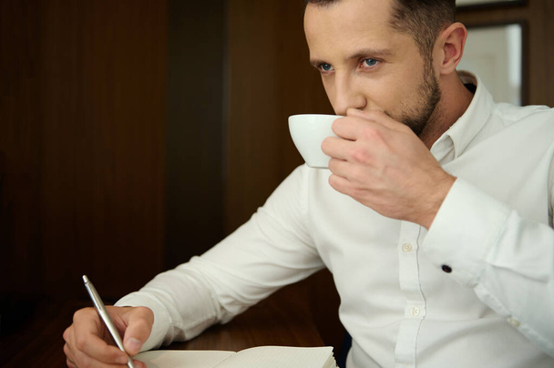 Close up portrait of a handsome attractive businessman, entrepreneur, journalist, writer holding silver pen and taking notes in notebook while drinking delicious freshly brewed coffee in his office - Photo, Image